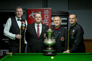 Peter Gilchrist, Stephen Harrison, Chris Ellis and David Causier all stand at the baulk end of the table before the final posing for a photo. The John Roberts Trophy is on the table just in front of them. Harrison is the marker for the final and Ellis is the referee.