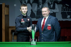 Antoni Kowalski shakes hands with tournament director Stuart Barker by the table. The trophy is on the table in between and in front of them.