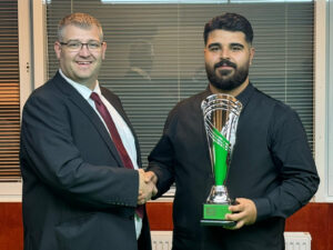 James Chambers the Tournament Director in suit and tie shakes Umut Dikme's hand as Dikme holds the trophy in his other hand.