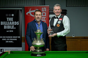 Paul Lloyd and Peter Gilchrist stand at the baulk end of the table posing for a photo. The John Roberts Trophy is on the table in front of Lloyd and Gilchrist is holding a smaller glass trophy which he gets to keep.